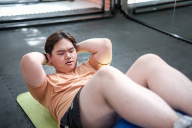 Spor salonunda desteklemek için spor topuyla egzersiz minderinde mekik çeken kilolu bir adam..