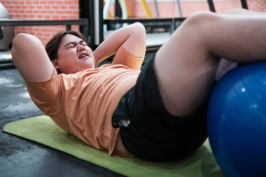 Spor salonunda desteklemek için spor topuyla egzersiz minderinde mekik çeken kilolu bir adam..