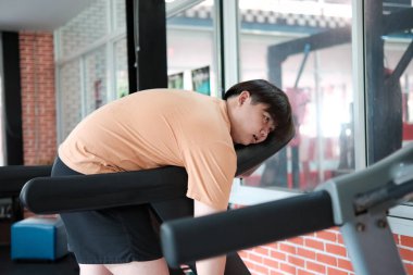 Kilolu bir adam koşu bandından eğilmiş, spor salonundaki antrenmandan sonra bitkin görünüyor..