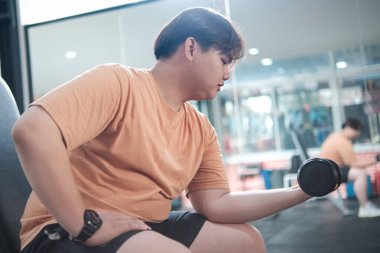 Kilolu bir adam bankta oturuyor, spor salonunda dambılı kaldırıyor..