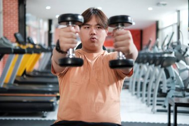 Kilolu bir adam spor salonunda önünde iki dambıl kaldırıyor..