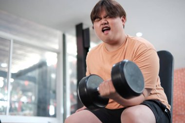 Kilolu bir adam bankta oturuyor, spor salonunda ağır bir halteri kaldırırken yüzünü buruşturuyor..