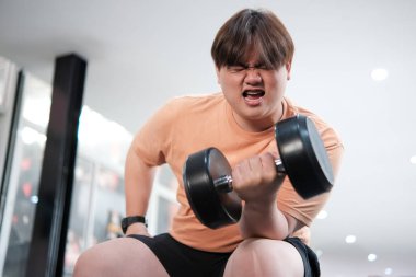 Kilolu bir adam bankta oturuyor, spor salonunda ağır bir halteri kaldırırken yüzünü buruşturuyor..