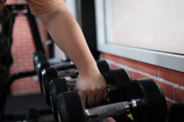 Bir kişi spor salonunda bir çift dambıl tutuyor. Dambıllar siyah ve üzerinde turuncu bir gömlek var.