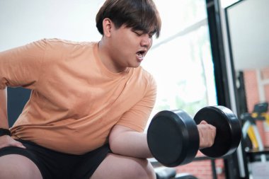 Turuncu gömlekli bir adam dambıl kaldırıyor. Spor salonundaki bir bankta oturuyor.