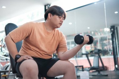 Turuncu gömlekli bir adam dambıl kaldırıyor. Spor salonundaki bir bankta oturuyor.