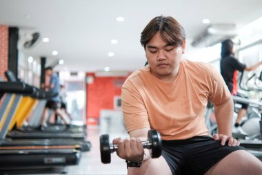 Turuncu gömlekli bir adam dambıl kaldırıyor. Spor salonundaki bir bankta oturuyor.