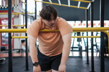 Bir adam, elleri sarı çubuğun üzerinde bir barda asılı duruyor. Terliyor ve yüzünde kaşları çatık.