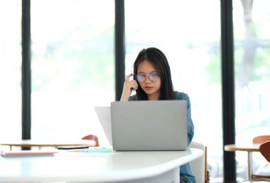 Önünde dizüstü bilgisayarı olan bir kadın masada oturuyor. Gözlük takıyor ve işine odaklanmış durumda. Sahne sessiz ve çalışkan bir atmosferi işaret ediyor.