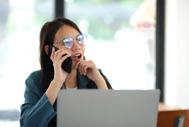 Bir kadın elinde dizüstü bilgisayarla bir masada otururken cep telefonuyla konuşuyor. Gözlük takıyor ve ciddi ya da odaklanmış durumda.
