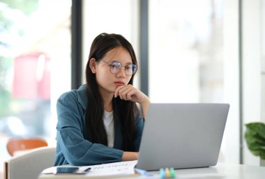 Önünde dizüstü bilgisayar olan bir kadın masada oturuyor. Gözlük takıyor ve derin düşüncelere dalmış.