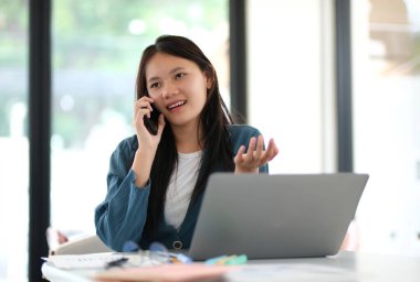 Bir kadın elinde dizüstü bilgisayarla bir masada otururken cep telefonuyla konuşuyor. Mavi bir ceket giyiyor ve profesyonel bir ortamda.