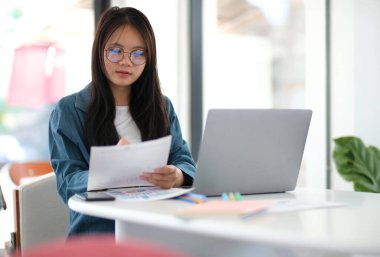 Bir kadın elinde dizüstü bilgisayar ve bir yığın kağıtla masada oturuyor. Gözlük takıyor ve işine odaklanmış durumda.