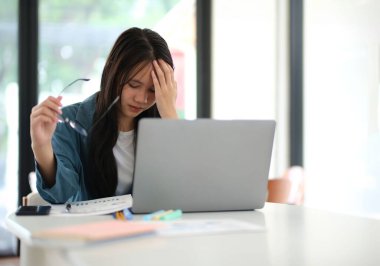 Masasında dizüstü bilgisayarı ve not defteri olan bir kadın oturuyor. Gözlük takıyor ve zor durumda.