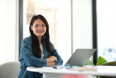 Önünde dizüstü bilgisayarı olan bir kadın masada oturuyor. Gülümsüyor ve keyfi yerinde.