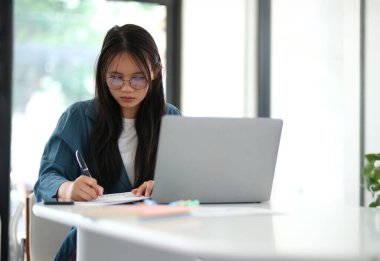 Bir kadın dizüstü bilgisayarı ve kalemi olan bir masada oturuyor. Bir kağıt parçasına yazıyor.