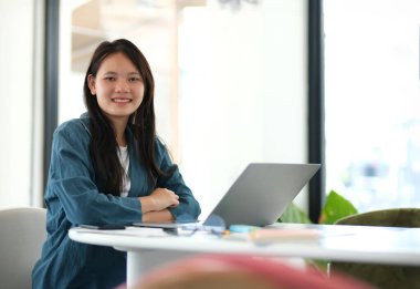 Önünde dizüstü bilgisayarı olan bir kadın masada oturuyor. Gülümsüyor ve mutlu.