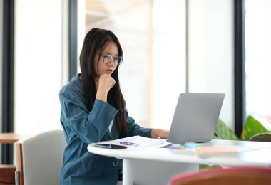 Önünde dizüstü bilgisayar olan bir kadın masada oturuyor. Gözlük takıyor ve işine odaklanmış durumda. Sahne profesyonel ya da akademik bir ortam öneriyor