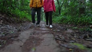 Ormanda kirli bir yolda yürüyen iki kişi. Biri sarı ceket giyiyor, diğeri de pembe ceket giyiyor.