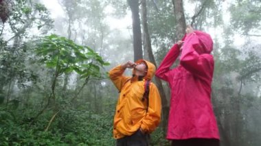 Yağmurluk giymiş iki kişi bir ağaca bakıyor. Bir tanesi bir şeyi işaret ediyor.