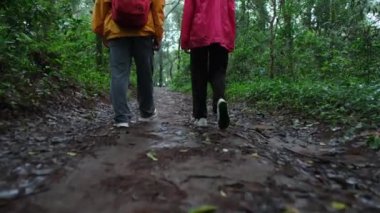 İki kişi ormanda çamurlu bir yolda yürüyor. İçlerinden biri sırt çantası takıyor.
