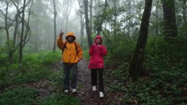 İki kişi ormanda dikiliyor, biri sarı ceketli, diğeri kırmızı ceketli.