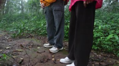 İki kişi ormanda dikiliyor, biri kırmızı gömlek giyiyor. Orman yemyeşil ve yemyeşil, bir sürü yaprak ve ağaçla dolu.