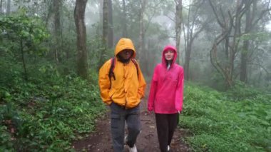 Bir adam ve bir kadın ormanda yürüyor, ikisi de yağmurluk giyiyor.