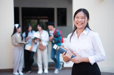 Kampüste elinde kitap tutan gülümseyen bir üniversite öğrencisi, arka planda diğer öğrencilerle birlikte güven ve pozitiflik yayıyor..