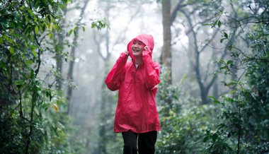 A smiling woman in a red raincoat hiking through a lush, misty forest on a rainy day, surrounded by greenery and enjoying the serene atmosphere and natural beauty. clipart