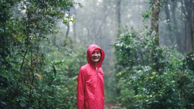 A smiling woman in a red raincoat hiking through a lush, misty forest on a rainy day, surrounded by greenery and enjoying the serene atmosphere and natural beauty. clipart