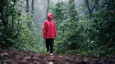 A smiling woman in a red raincoat hiking through a lush, misty forest on a rainy day, surrounded by greenery and enjoying the serene atmosphere and natural beauty. clipart