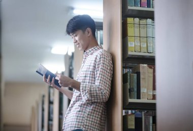 Bir üniversite öğrencisi, kitaplarla çevrili bir kitap okumaya dalmış bir şekilde kütüphanede duruyor..
