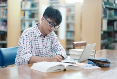 Genç Asyalı erkek üniversite öğrencisi kütüphanede tek başına araştırma yapıyor. Genç adam oturuyor ve bir proje üzerinde çalışıyor. Odaklanmış adam okuma ve yazma.