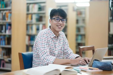 Genç Asyalı erkek üniversite öğrencisi kütüphanede tek başına araştırma yapıyor. Genç adam oturuyor ve bir proje üzerinde çalışıyor. Odaklanmış adam okuma ve yazma.