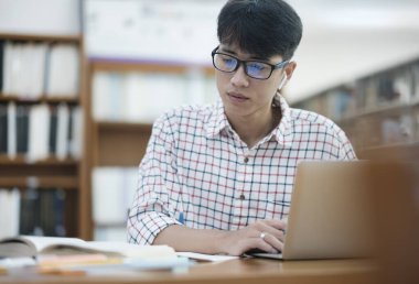 Genç Asyalı erkek üniversite öğrencisi kütüphanede tek başına araştırma yapıyor. Genç adam oturuyor ve bir proje üzerinde çalışıyor. Odaklanmış adam okuma ve yazma.