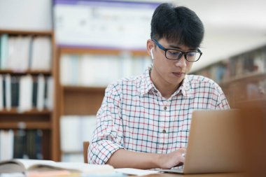 Genç Asyalı erkek üniversite öğrencisi kütüphanede tek başına araştırma yapıyor. Genç adam oturuyor ve bir proje üzerinde çalışıyor. Odaklanmış adam okuma ve yazma.