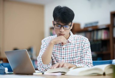 Genç Asyalı erkek üniversite öğrencisi kütüphanede tek başına araştırma yapıyor. Genç adam oturuyor ve bir proje üzerinde çalışıyor. Odaklanmış adam okuma ve yazma.