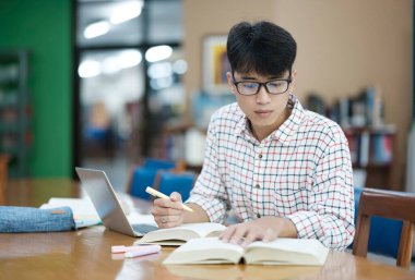 Genç Asyalı erkek üniversite öğrencisi kütüphanede tek başına araştırma yapıyor. Genç adam oturuyor ve bir proje üzerinde çalışıyor. Odaklanmış adam okuma ve yazma.
