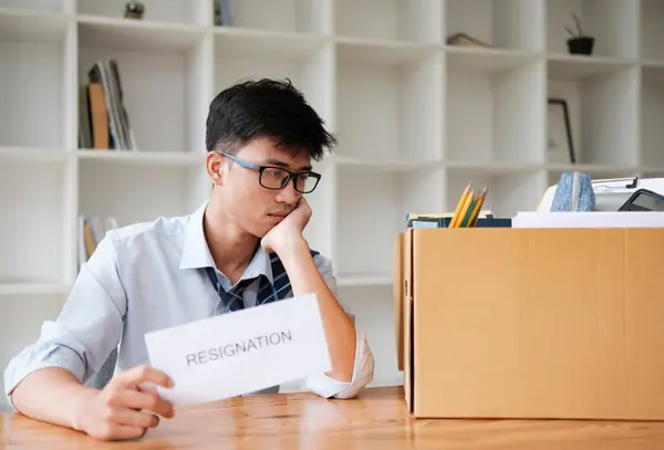 stock image A businessman looks thoughtful while holding a resignation letter, with a box of personal belongings beside him. The image symbolizes job resignation, career change, or leaving a position.