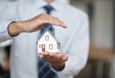 Bir emlakçı bir ev modelini elinde tutarken tek eliyle onu koruyor, gayrimenkul, ev sahipliği ve sigorta korumasını temsil ediyor..