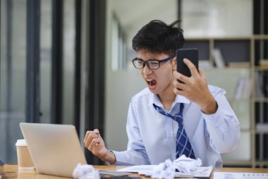 Sinirli iş adamı masasında çalışırken öfke ve stresi ifade ediyor. Bilgisayarına bağırıyor, iş sorunları, iş stresi ya da proje başarısızlığı yüzünden buruşuk kağıtlarla çevrili..