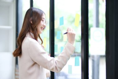 Bir iş kadını yapışkan notlara yazı yazarken görülüyor, cam bir duvara iş stratejisi planlarken. Görüntü odak, yaratıcılık ve stratejik düşünceyi temsil ediyor. Liderlik ve kararın altını çiziyor
