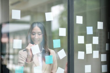 Bir iş kadını yapışkan notlara yazı yazarken görülüyor, cam bir duvara iş stratejisi planlarken. Görüntü odak, yaratıcılık ve stratejik düşünceyi temsil ediyor. Liderlik ve kararın altını çiziyor