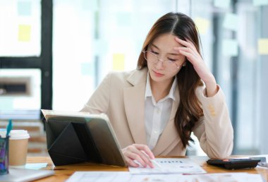 Profesyonel bir iş kadını, masasındaki tablet ve yazılı çizelgelere bakarken derin bir şekilde odaklanır. Modern ofis ortamında kahve, kırtasiye ve doğal ışıklandırma vardır..