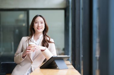 Neşeli bir iş kadını bir kahve fincanı tutuyor ve işe ara verirken gülümsüyor. Görüntü, arka planda modern teknolojiyle rahatlama, üretkenlik ve profesyonelliği aktarıyor..