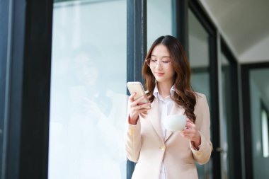 Bir iş kadını kahve fincanı tutarken gülümser ve mola sırasında telefonunu kontrol eder. Görüntü, dengeli bir çalışma ortamında modern profesyonelliği, üretkenliği ve rahatlamayı yansıtıyor..