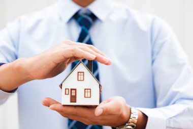 Bir emlakçı ev modelini bir eliyle tutarken diğer eliyle onu koruyor. Mülk korumasını, ev mülkiyetini ve emlak güvenliğini sembolize ediyor..