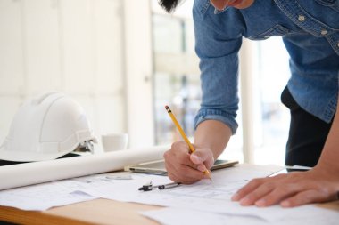 Evin planlarını çizmek için kalem kullanan genç bir mimarın yakın çekimi. Masa bir pusula, tablet ve güvenlik kaskı gibi tasarım araçları içerir. Doğal ışık alan parlak ve açık ofis alanı.