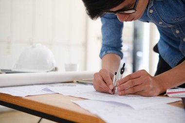 Evin planlarını çizmek için pusula kullanan bir mimarın eli. Masanın tablet, kalem ve güvenlik kaskı gibi tasarım araçları var. Doğal ışıklandırmalı parlak ofis alanı.